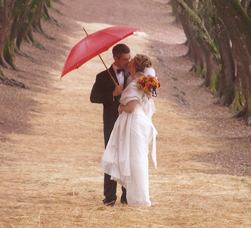 Wedding photo of Hugh and Monique Davies kissing under a red umbrella in the olive grove.